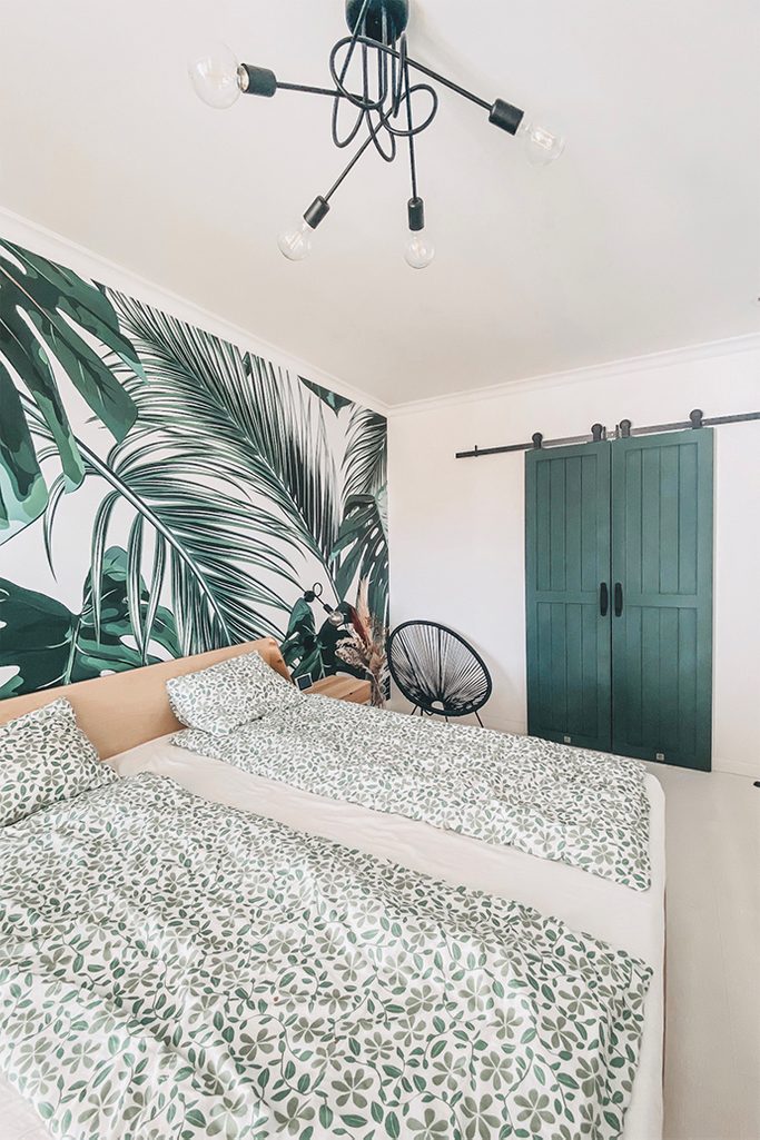 green doors, bedroom with walk-in wardrobe, sliding door to the wardrobe, sliding door in the bedroom, greenery in the bedroom, industrial chandelier in the bedroom, decorative wallpaper with plants, botanical wallpaper, green wallpaper for the bedroom, openwork chair, plant motif on bedding, green color bedroom