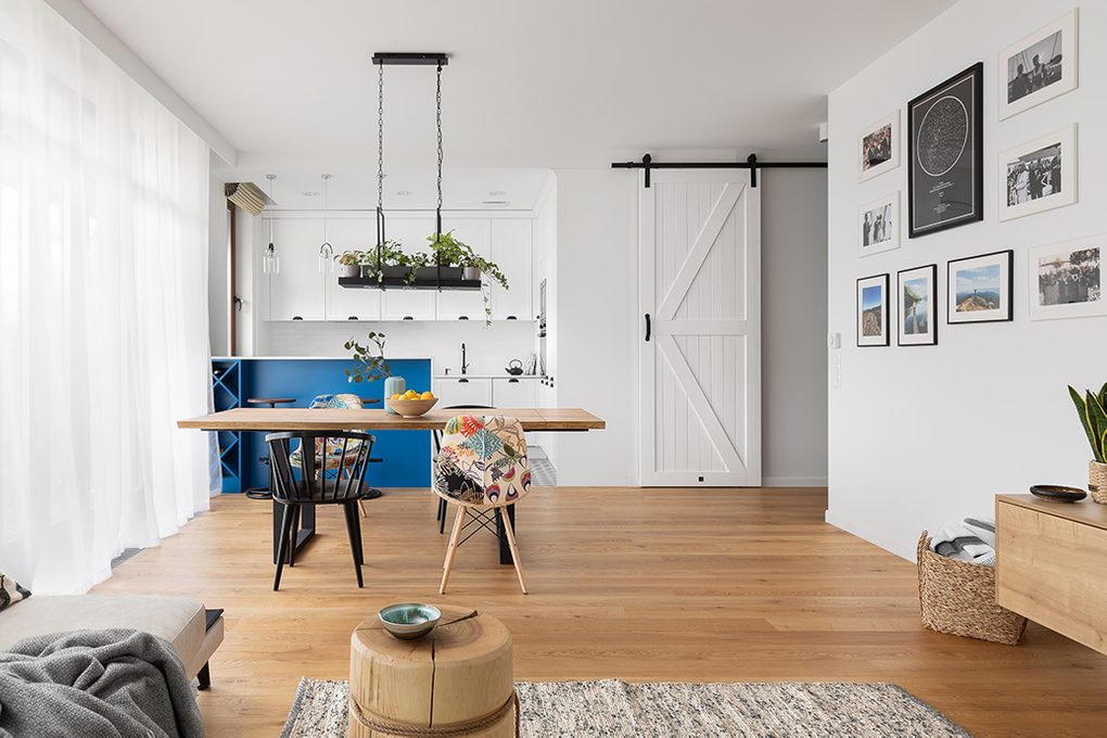 bright living room with dining table and open kitchen, white kitchen separated with breakfast bar worktop painted in blue, white sliding door to the utility room, photo gallery on the wall